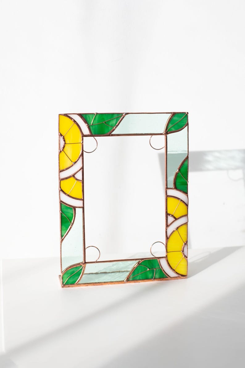 May include: A rectangular stained glass mirror with a vibrant design. The frame features yellow, green, and white geometric patterns, resembling lemons and leaves. The mirror is supported by a copper-colored base.