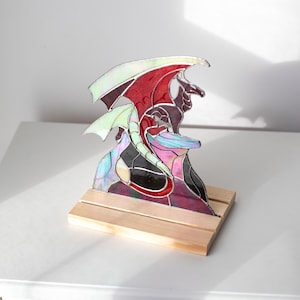 Op de afbeelding: Gebrandschilderde glazen drakenbeeldhouwwerk met rode, witte en iriserende vleugels, op een houten voet. De draak heeft een gedetailleerd ontwerp met een zwart lichaam en paarse accenten. Een uniek decoratief stuk.