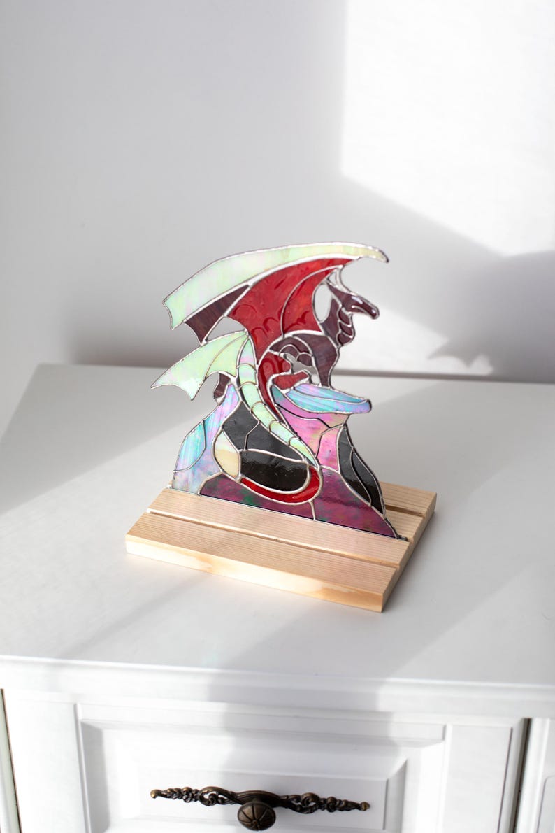 Op de afbeelding: Gebrandschilderde glazen drakenbeeldhouwwerk met rode, witte en iriserende vleugels, op een houten voet. De draak heeft een gedetailleerd ontwerp met een zwart lichaam en paarse accenten. Een uniek decoratief stuk.
