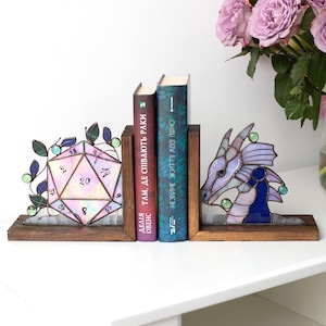 May include: Stained glass bookends featuring a D20 die and a dragon head design. The bookends are set on a wooden base, with books between them. The die is pink and purple, and the dragon is purple and blue.
