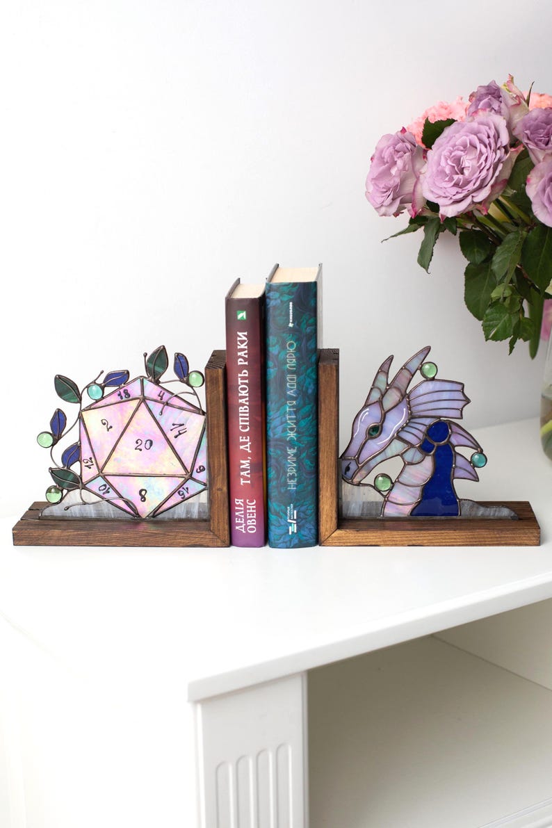 May include: Stained glass bookends featuring a D20 die and a dragon head design. The bookends are set on a wooden base, with books between them. The die is pink and purple, and the dragon is purple and blue.
