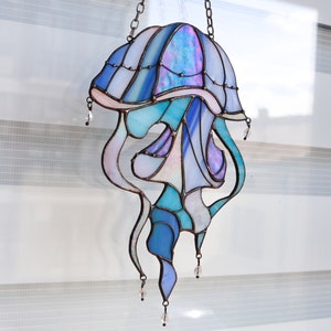 Op de afbeelding: Een gebrandschilderd glas-zonnevanger in de vorm van een kwal met iriserende blauwe, paarse en roze glazen. De kwal hangt aan een ketting en heeft kristallen kralen die aan de onderkant hangen.