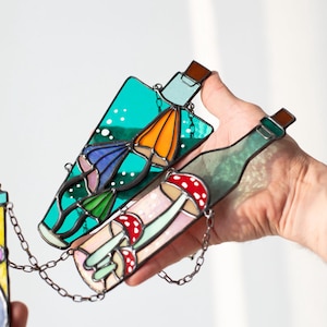 May include: Two stained glass bottles hanging from a chain. One bottle is teal blue with a white background and features a mushroom with a red cap and a blue and orange mushroom. The other bottle is green with a white background and features a mushroom with a red cap and a white stem.