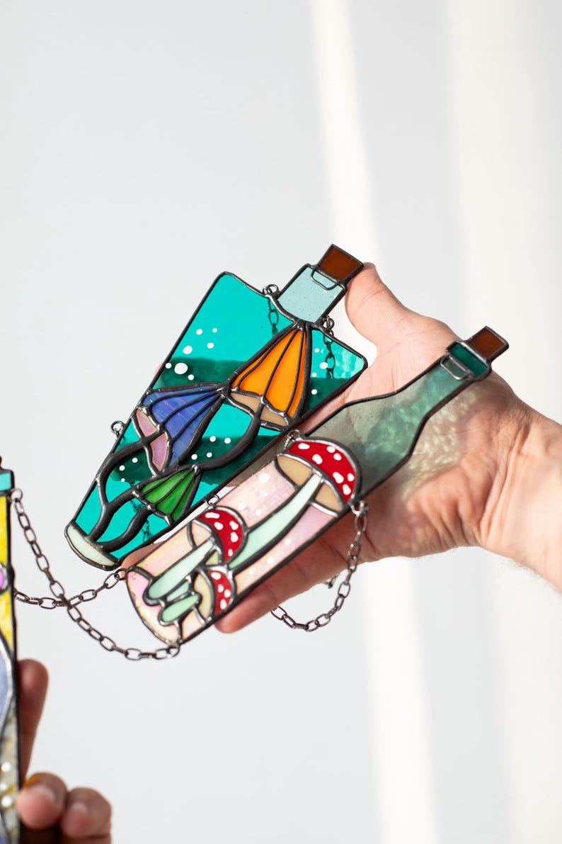 May include: Two stained glass bottles hanging from a chain. One bottle is teal blue with a white background and features a mushroom with a red cap and a blue and orange mushroom. The other bottle is green with a white background and features a mushroom with a red cap and a white stem.