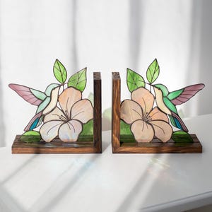 Pu&ograve; includere: Un paio di fermalibri in legno con disegni di colibr&igrave; e fiori in vetro colorato. I colibr&igrave; hanno ali rosa, verdi e blu. I fiori sono color pesca con foglie verdi. I fermalibri sono su una superficie bianca.
