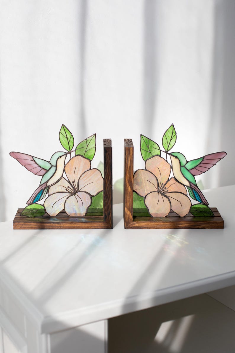 Pu&ograve; includere: Un paio di fermalibri in legno con disegni di colibr&igrave; e fiori in vetro colorato. I colibr&igrave; hanno ali rosa, verdi e blu. I fiori sono color pesca con foglie verdi. I fermalibri sono su una superficie bianca.
