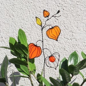 K&ouml;nnte beinhalten: Eine Drahtskulptur einer Pflanze mit orangefarbenen und gelben Glasblumen. Die Skulptur steht vor einer gr&uuml;nen, belaubten Pflanze.
