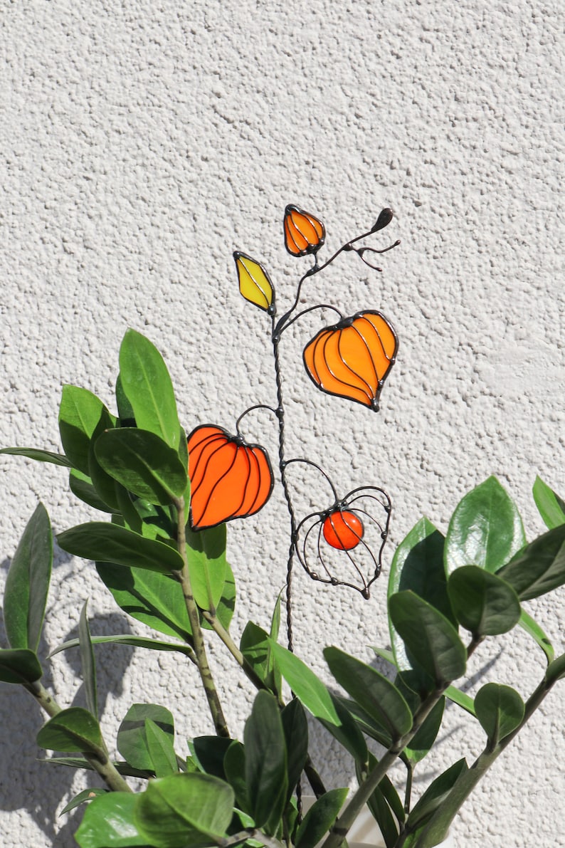 K&ouml;nnte beinhalten: Eine Drahtskulptur einer Pflanze mit orangefarbenen und gelben Glasblumen. Die Skulptur steht vor einer gr&uuml;nen, belaubten Pflanze.