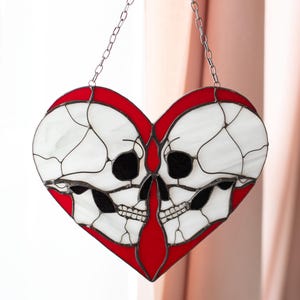 May include: Heart-shaped stained glass suncatcher with a design of two skulls. The skulls are white with black eye sockets and teeth, set against a red heart outline. A silver chain is attached for hanging. Gothic home decor.