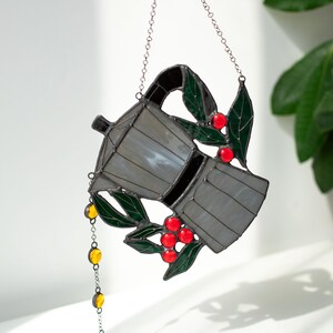 Puede incluir: Un atrapasoles de vidrieras con forma de cafetera moka, con paneles de vidrio gris, hojas de color verde oscuro y bayas de color rojo brillante. Una peque&ntilde;a cadena con cuentas de vidrio amarillas cuelga del pico, suspendida por una cadena de metal plateado.