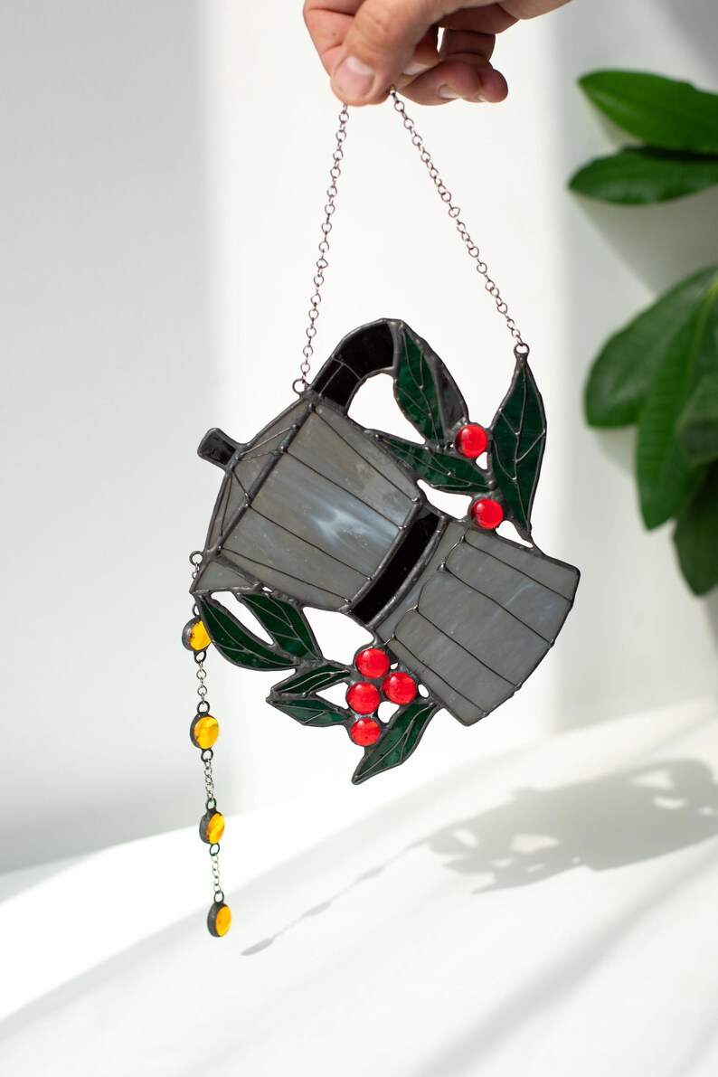 Puede incluir: Un atrapasoles de vidrieras con forma de cafetera moka, con paneles de vidrio gris, hojas de color verde oscuro y bayas de color rojo brillante. Una peque&ntilde;a cadena con cuentas de vidrio amarillas cuelga del pico, suspendida por una cadena de metal plateado.