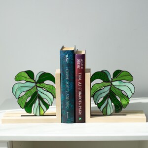 K&ouml;nnte beinhalten: Dekorative Buchst&uuml;tzen mit einem Design aus Monstera-Bl&auml;ttern aus Glas in Gr&uuml;nt&ouml;nen. Die h&ouml;lzernen Buchst&uuml;tzen halten zwei B&uuml;cher mit blauen und violetten Einb&auml;nden. Die B&uuml;cher haben Text in einer Fremdsprache.