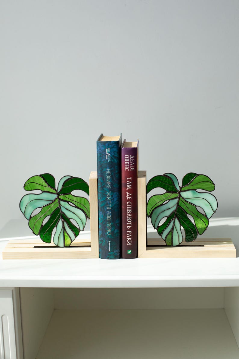 K&ouml;nnte beinhalten: Dekorative Buchst&uuml;tzen mit einem Design aus Monstera-Bl&auml;ttern aus Glas in Gr&uuml;nt&ouml;nen. Die h&ouml;lzernen Buchst&uuml;tzen halten zwei B&uuml;cher mit blauen und violetten Einb&auml;nden. Die B&uuml;cher haben Text in einer Fremdsprache.