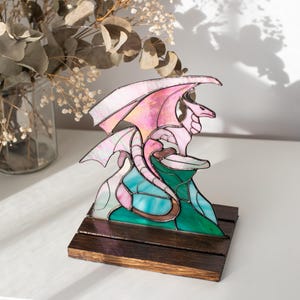 Op de afbeelding: Een gebrandschilderde drakenbeeldhouwwerk met roze en witte vleugels, bovenop een turquoise en groene berg. De draak staat op een donkerbruine houten basis. De sculptuur wordt op een wit oppervlak tentoongesteld.