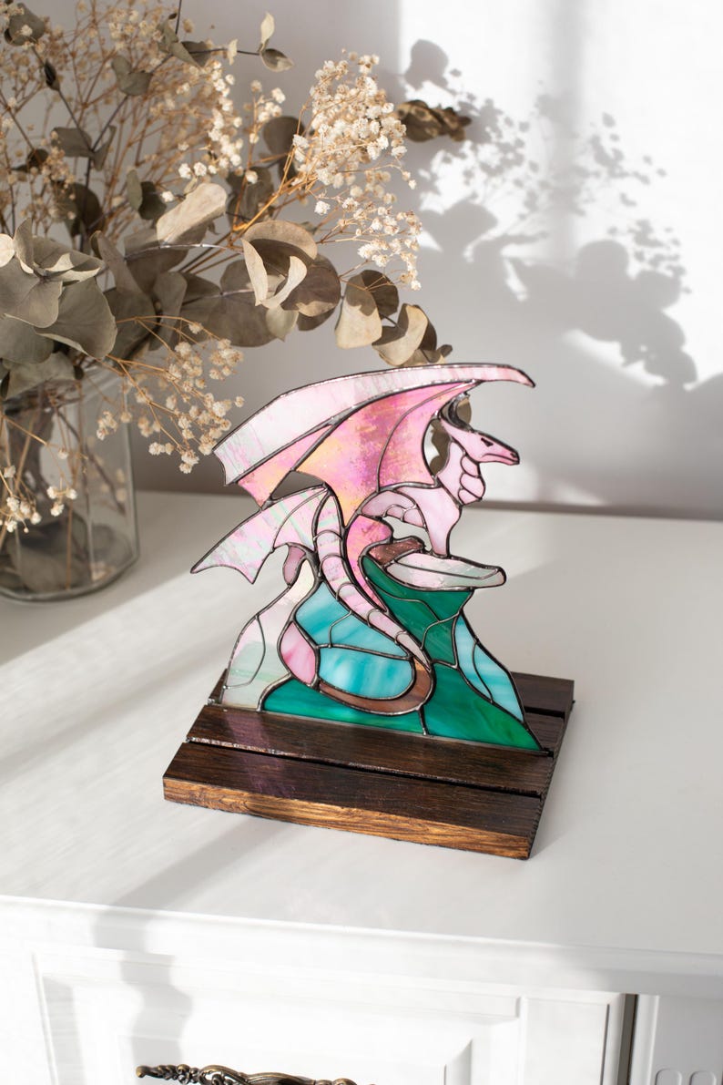 Op de afbeelding: Een gebrandschilderde drakenbeeldhouwwerk met roze en witte vleugels, bovenop een turquoise en groene berg. De draak staat op een donkerbruine houten basis. De sculptuur wordt op een wit oppervlak tentoongesteld.