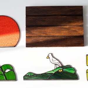 Op de afbeelding: Een verzameling glas-in-lood kunstwerken. Bevat een rode en oranje zon, een donker houten paneel, groen gebladerte, een vogel op een alligator en gele en groene bladeren. Elk stuk heeft een zwarte rand.