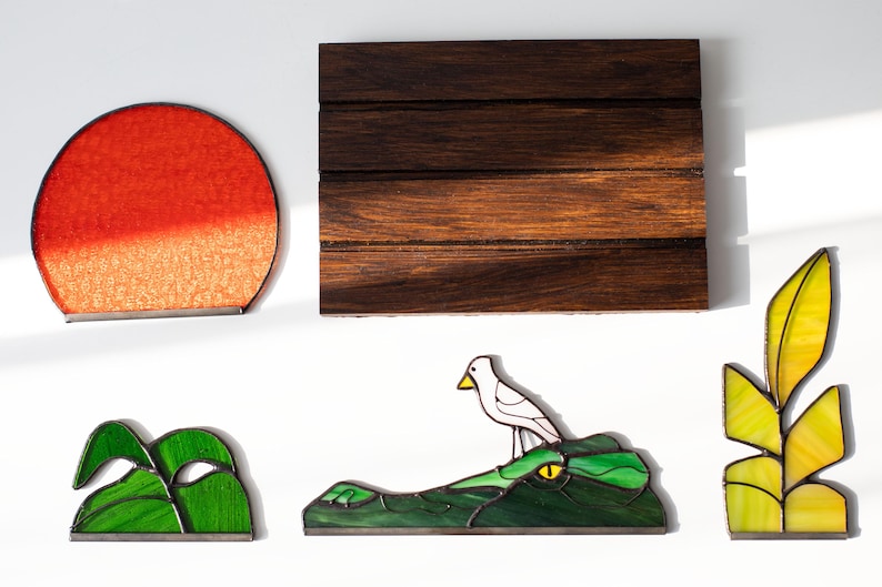 Op de afbeelding: Een verzameling glas-in-lood kunstwerken. Bevat een rode en oranje zon, een donker houten paneel, groen gebladerte, een vogel op een alligator en gele en groene bladeren. Elk stuk heeft een zwarte rand.