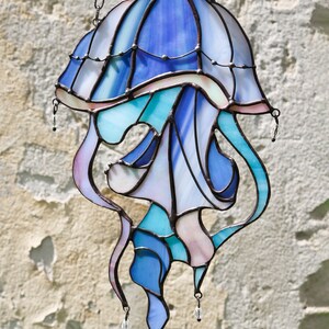 Op de afbeelding: Een glas-in-lood kwal hangend aan een ketting. De kwal is blauw, paars en roze met heldere glazen accenten. De kwal heeft een klokvormige bovenkant en lange, vloeiende tentakels.
