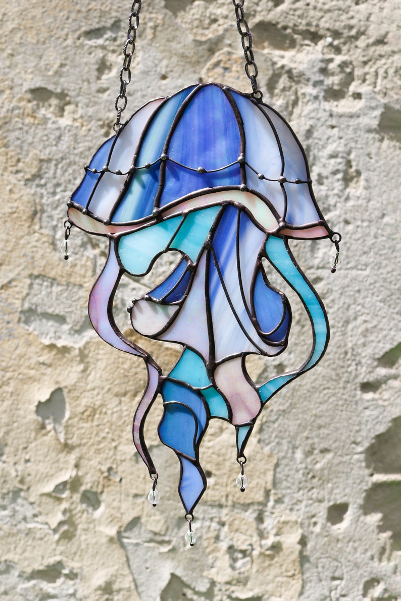 Op de afbeelding: Een glas-in-lood kwal hangend aan een ketting. De kwal is blauw, paars en roze met heldere glazen accenten. De kwal heeft een klokvormige bovenkant en lange, vloeiende tentakels.