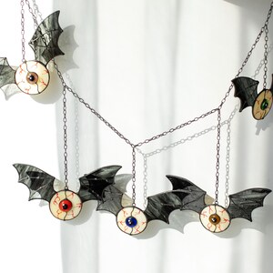 May include: A hanging mobile featuring five bat-shaped ornaments connected by dark metal chains. Each bat has black stained glass wings and a central circular body resembling a human eyeball with a coloured iris in red, blue, green, or brown.