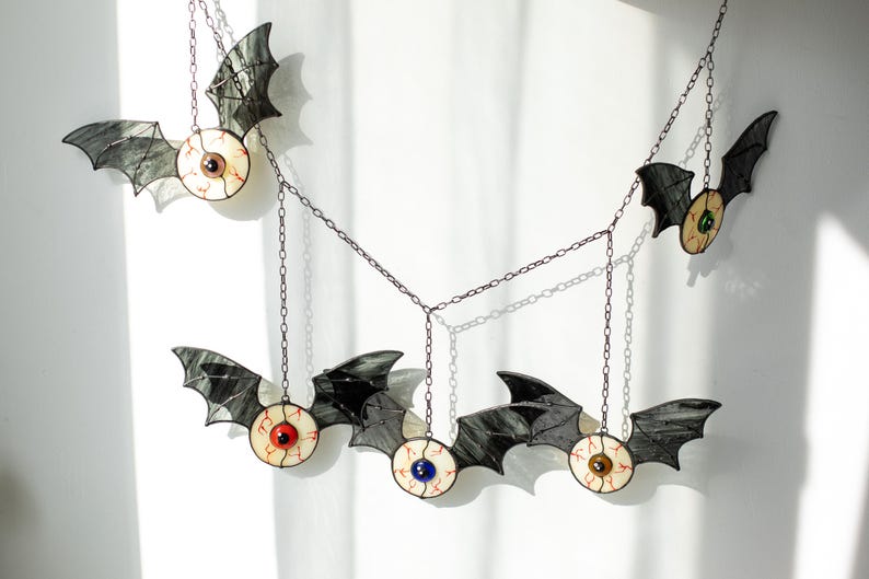 May include: A hanging mobile featuring five bat-shaped ornaments connected by dark metal chains. Each bat has black stained glass wings and a central circular body resembling a human eyeball with a coloured iris in red, blue, green, or brown.