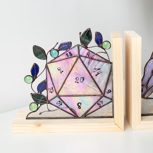 May include: A stained glass D20 die bookend with a wooden base. The die is iridescent with pink, purple, and green hues, and features black numbers. Green and blue glass leaves and beads adorn the die. The bookend is a unique decorative item.