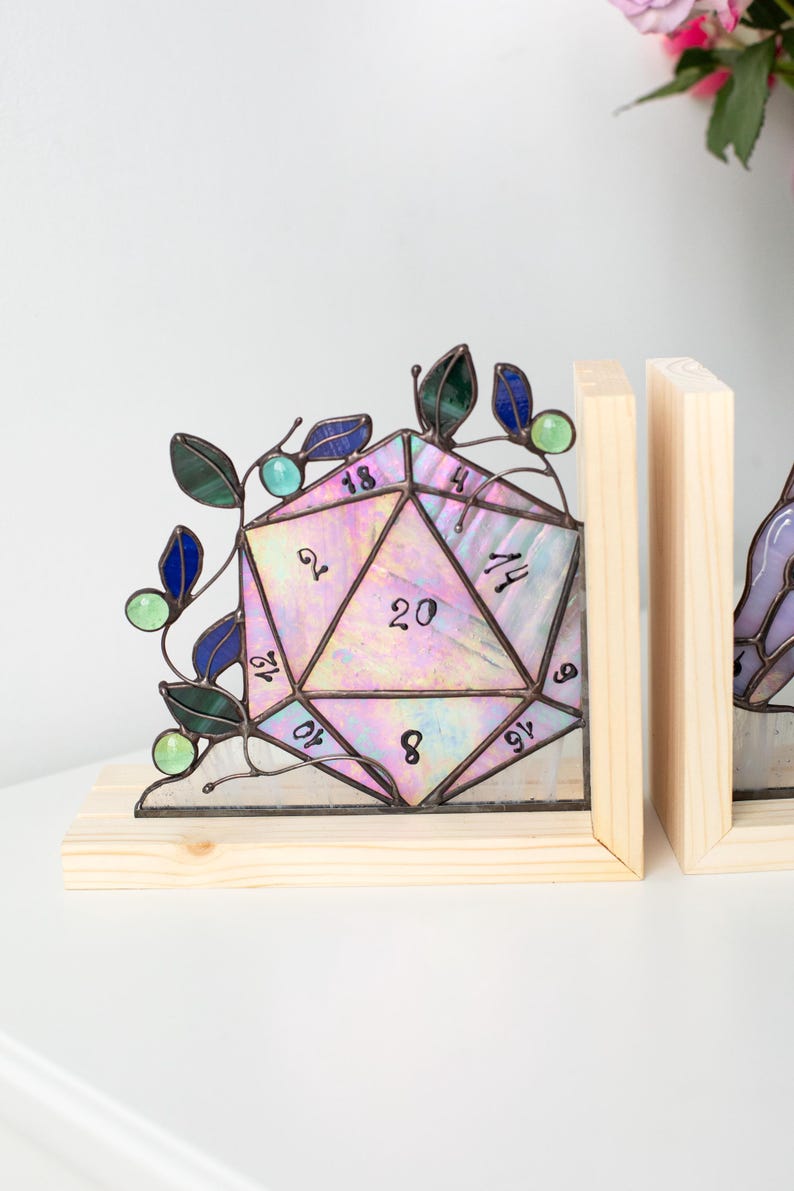 May include: A stained glass D20 die bookend with a wooden base. The die is iridescent with pink, purple, and green hues, and features black numbers. Green and blue glass leaves and beads adorn the die. The bookend is a unique decorative item.
