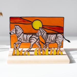 以下が含まれることがあります： 黒と白の縞模様の2頭のシマウマが、オレンジ色の夕焼けと黄色の太陽を背景にしたステンドグラスのアート作品。この作品は木の台座に取り付けられており、サバンナのシーンを表現しています。