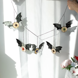 May include: A hanging mobile featuring five stained glass bats, each with a central realistic eyeball design in various colours including red, green, and blue. The bats are connected by thin metal chains, creating a decorative wall ornament.