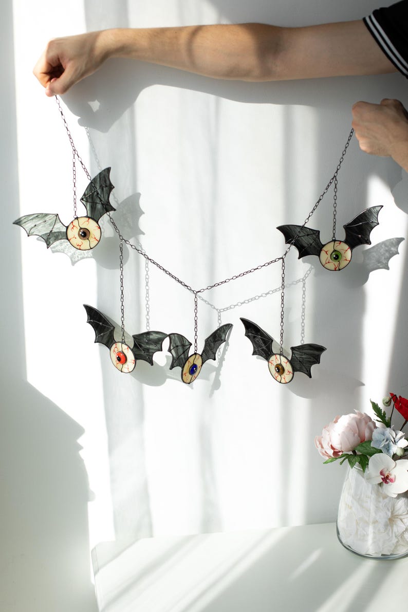 May include: A hanging mobile featuring five stained glass bats, each with a central realistic eyeball design in various colours including red, green, and blue. The bats are connected by thin metal chains, creating a decorative wall ornament.