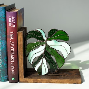 K&ouml;nnte beinhalten: Ein Buchst&uuml;tze aus Buntglas in Form eines Monstera-Blattes, in Gr&uuml;n- und Wei&szlig;t&ouml;nen. Sie steht auf einem dunklen Holzsockel, neben B&uuml;chern mit fremdsprachigem Text.