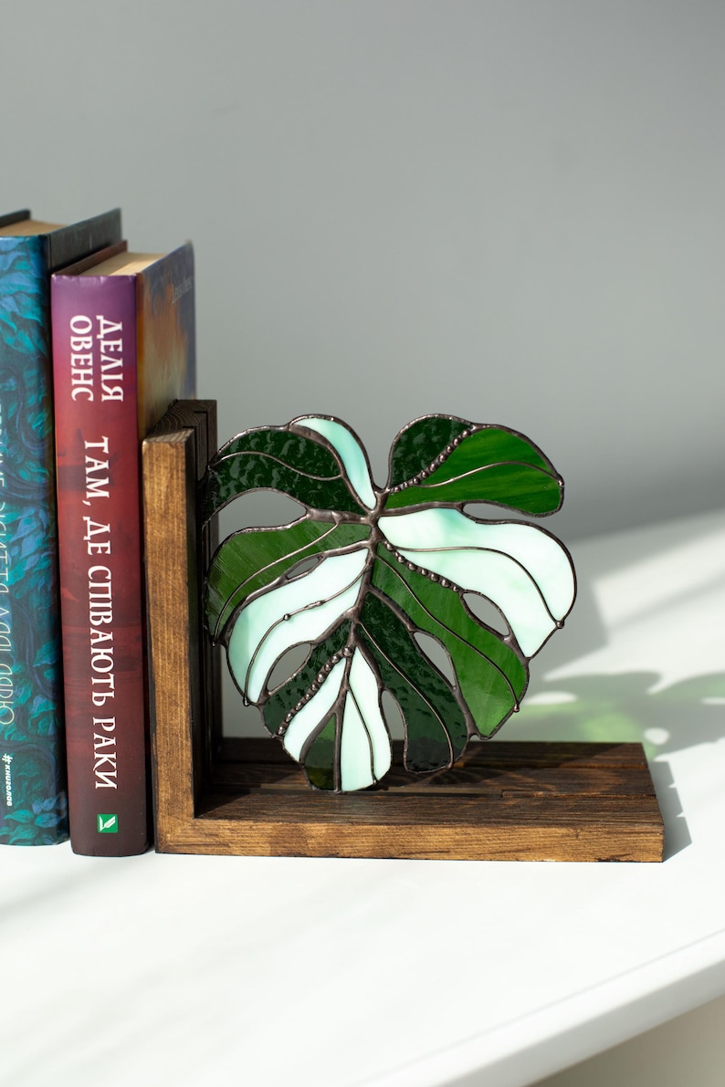 K&ouml;nnte beinhalten: Ein Buchst&uuml;tze aus Buntglas in Form eines Monstera-Blattes, in Gr&uuml;n- und Wei&szlig;t&ouml;nen. Sie steht auf einem dunklen Holzsockel, neben B&uuml;chern mit fremdsprachigem Text.