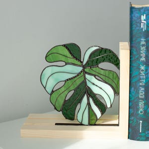 K&ouml;nnte beinhalten: Ein Buntglas-Monstera-Blatt in Gr&uuml;n- und Wei&szlig;t&ouml;nen, das in einem h&ouml;lzernen Buchst&uuml;tzen platziert ist. Das Blatt hat ein detailliertes Design mit sichtbaren Adern und einem strukturierten Aussehen. Ein Buch mit blauem Einband ist sichtbar.