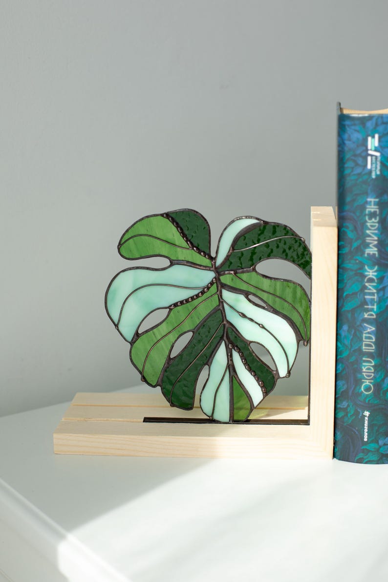 K&ouml;nnte beinhalten: Ein Buntglas-Monstera-Blatt in Gr&uuml;n- und Wei&szlig;t&ouml;nen, das in einem h&ouml;lzernen Buchst&uuml;tzen platziert ist. Das Blatt hat ein detailliertes Design mit sichtbaren Adern und einem strukturierten Aussehen. Ein Buch mit blauem Einband ist sichtbar.