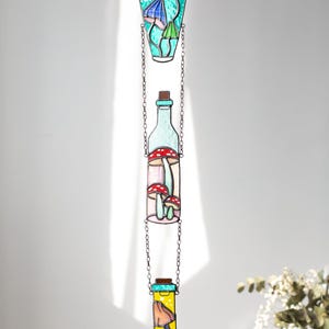 May include: Three stained glass bottles hanging from a chain. The top bottle is teal blue with yellow and blue mushrooms inside. The middle bottle is clear with red and white mushrooms inside. The bottom bottle is yellow with a blue mushroom inside.