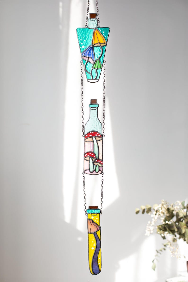 May include: Three stained glass bottles hanging from a chain. The top bottle is teal blue with yellow and blue mushrooms inside. The middle bottle is clear with red and white mushrooms inside. The bottom bottle is yellow with a blue mushroom inside.