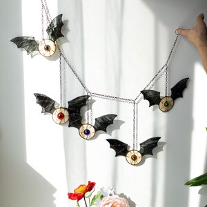 May include: A hanging mobile featuring five bat-shaped ornaments with stained glass wings and central eyeball designs. Each bat has a unique iris colour, including red, blue, green, brown, and dark brown, connected by thin metal chains for display.