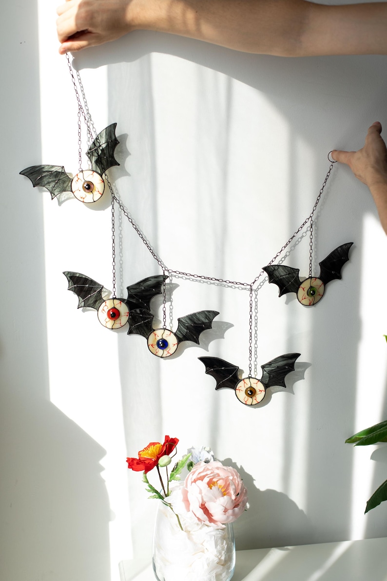 May include: A hanging mobile featuring five bat-shaped ornaments with stained glass wings and central eyeball designs. Each bat has a unique iris colour, including red, blue, green, brown, and dark brown, connected by thin metal chains for display.