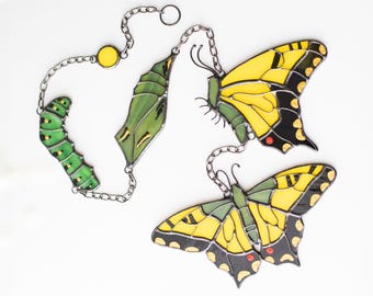 Puede incluir: Un móvil de mariposa de vidrio coloreado con una oruga, una crisálida y dos mariposas. Las mariposas son amarillas con marcas negras y rojas. El móvil está suspendido de una cadena plateada.