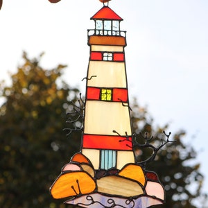 May include: A stained glass lighthouse with red and white stripes, a yellow window, and a blue door. The lighthouse is on a rocky base with orange and pink rocks and blue waves.