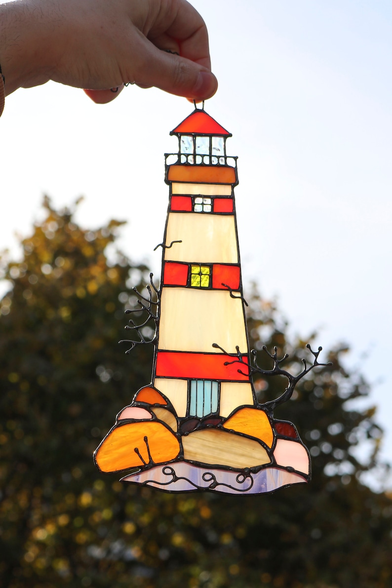 May include: A stained glass lighthouse with red and white stripes, a yellow window, and a blue door. The lighthouse is on a rocky base with orange and pink rocks and blue waves.