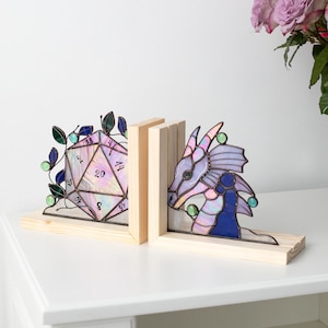 May include: Stained glass bookends featuring a D20 die and a dragon head. The bookends are set on a light wood base. The dragon is primarily purple and blue with green accents. The die is a light pink with black numbers.