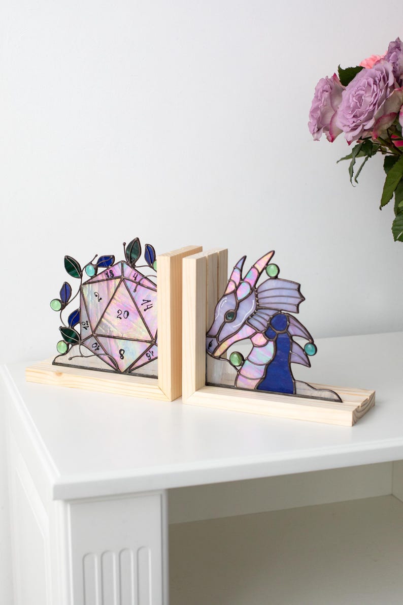May include: Stained glass bookends featuring a D20 die and a dragon head. The bookends are set on a light wood base. The dragon is primarily purple and blue with green accents. The die is a light pink with black numbers.