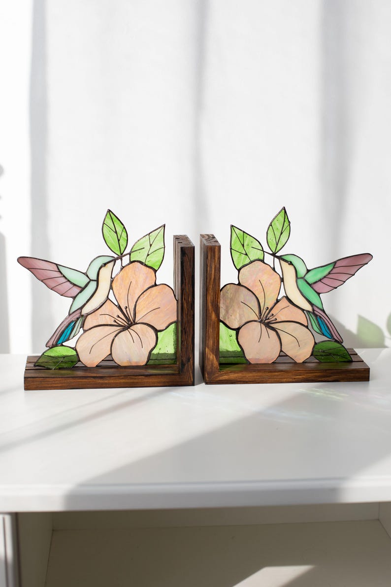 Pu&ograve; includere: Un paio di fermalibri in legno con un design a vetrate. Ogni fermalibri presenta un colibr&igrave;, un grande fiore e foglie verdi. Il colibr&igrave; ha ali rosa, blu e verdi. Il fiore &egrave; color pesca.