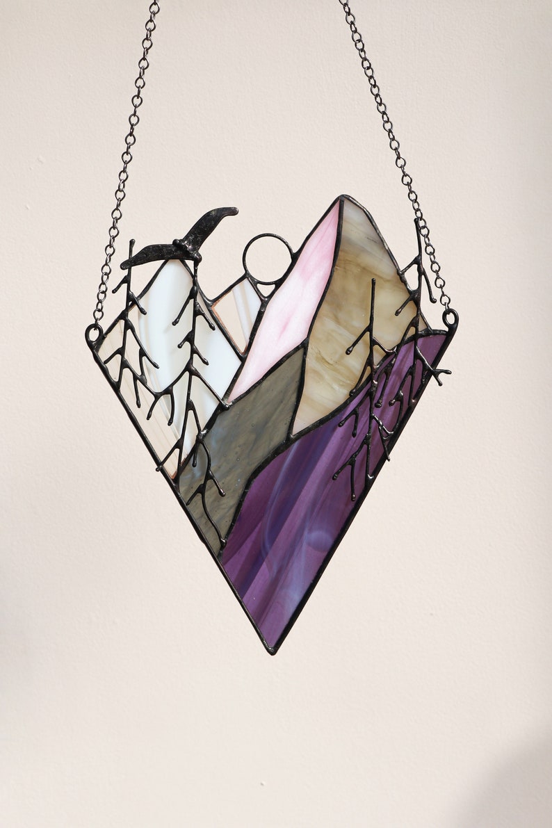 May include: A stained glass window hanging from a chain, depicting a purple, pink, and brown mountain range with a black bird flying overhead.