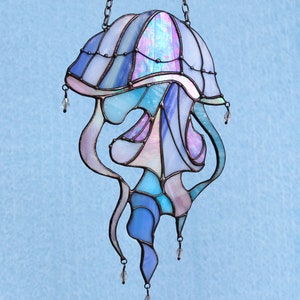 Op de afbeelding: Een gebrandschilderd glas-zonnevanger in de vorm van een kwal met iriserende blauwe, paarse en roze glasstukken. De kwal hangt aan een zwarte ketting met kristallen kralen.