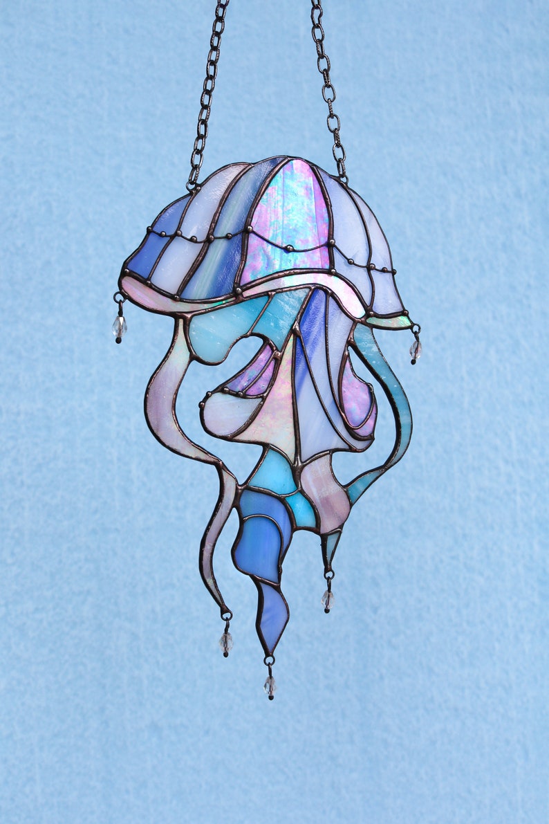 Op de afbeelding: Een gebrandschilderd glas-zonnevanger in de vorm van een kwal met iriserende blauwe, paarse en roze glasstukken. De kwal hangt aan een zwarte ketting met kristallen kralen.