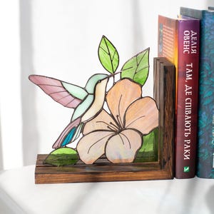 Pu&ograve; includere: Reggilibri con colibr&igrave; e fiori in vetro colorato. Il colibr&igrave; ha accenti rosa, blu e verdi. Il fiore &egrave; color pesca con una base in legno marrone scuro. I reggilibri sono accanto a una fila di libri.