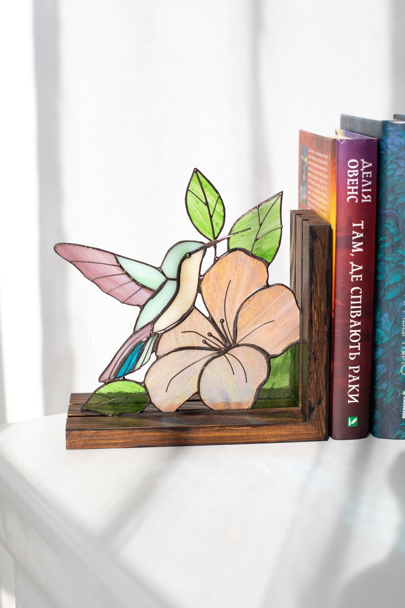 Pu&ograve; includere: Reggilibri con colibr&igrave; e fiori in vetro colorato. Il colibr&igrave; ha accenti rosa, blu e verdi. Il fiore &egrave; color pesca con una base in legno marrone scuro. I reggilibri sono accanto a una fila di libri.