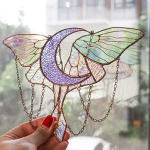 May include: A stained glass window hanging with a crescent moon and iridescent butterfly wings. The wings are green, purple, and white. The moon is purple and the hanging chains are copper.